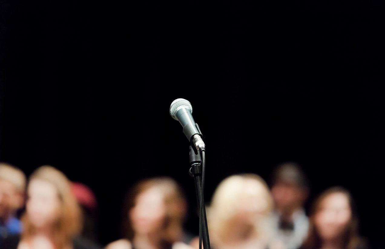 A wired silver microphone placed on a black mic stand in front of a blurry audience